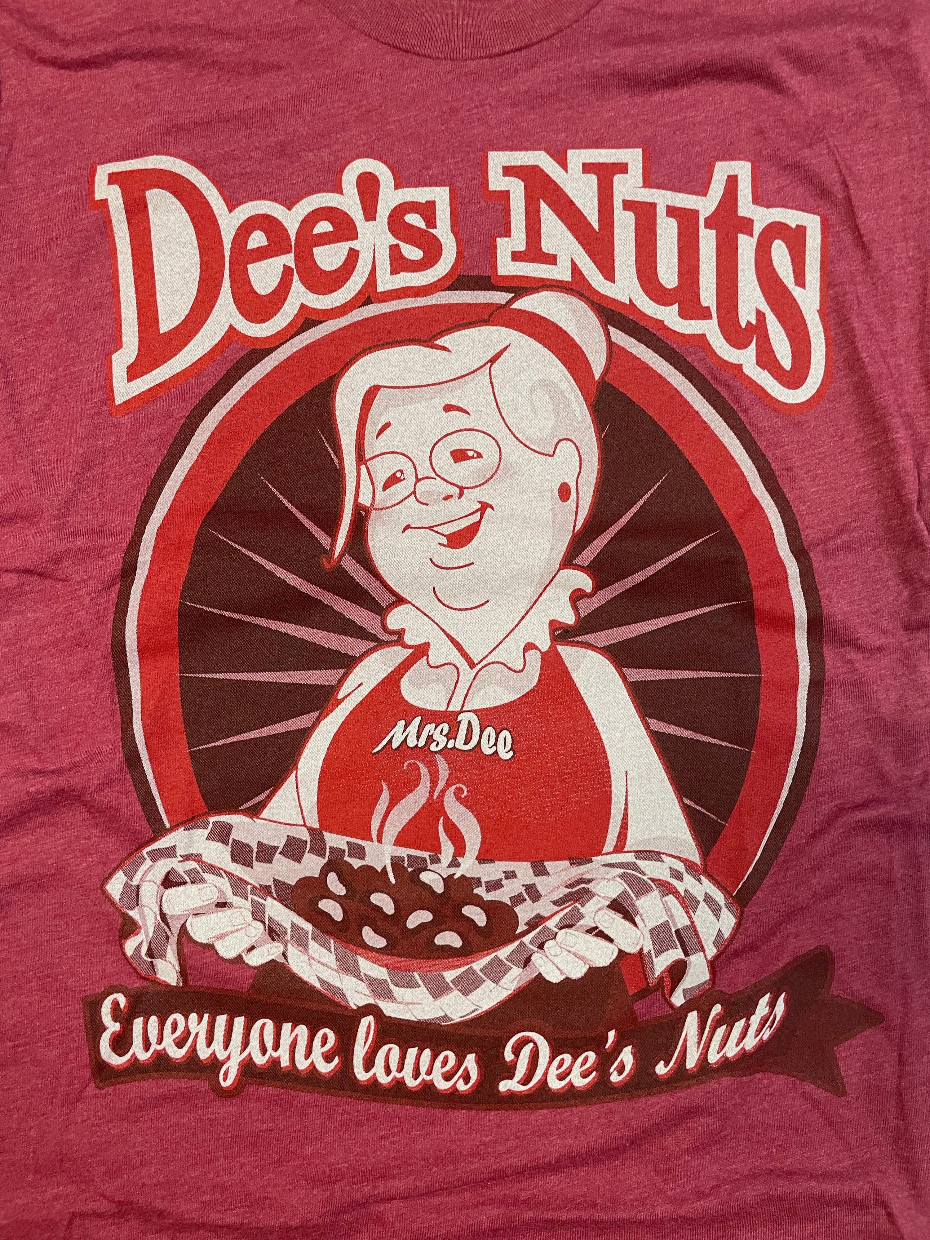 Close-up of a red Dee's Nuts t-shirt featuring a cartoon character named Mrs. Dee holding a steaming tray of peanuts with a checkered cloth. The design includes the bold text “Dee’s Nuts” at the top and the slogan “Everyone loves Dee’s Nuts” on a banner at the bottom. Playful vintage-style branding promoting the snack company.