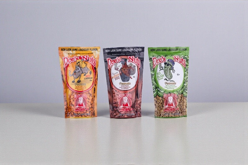 Three Dee’s Nuts flavored peanut bags standing side by side on a neutral background, featuring different colorful packaging designs and visible peanuts inside each resealable pouch.