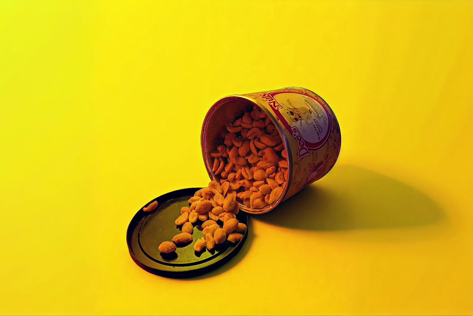 A can of Dee's Nuts flavored peanuts is tipped over on a bright yellow background, with peanuts spilling out onto the black lid beneath it.